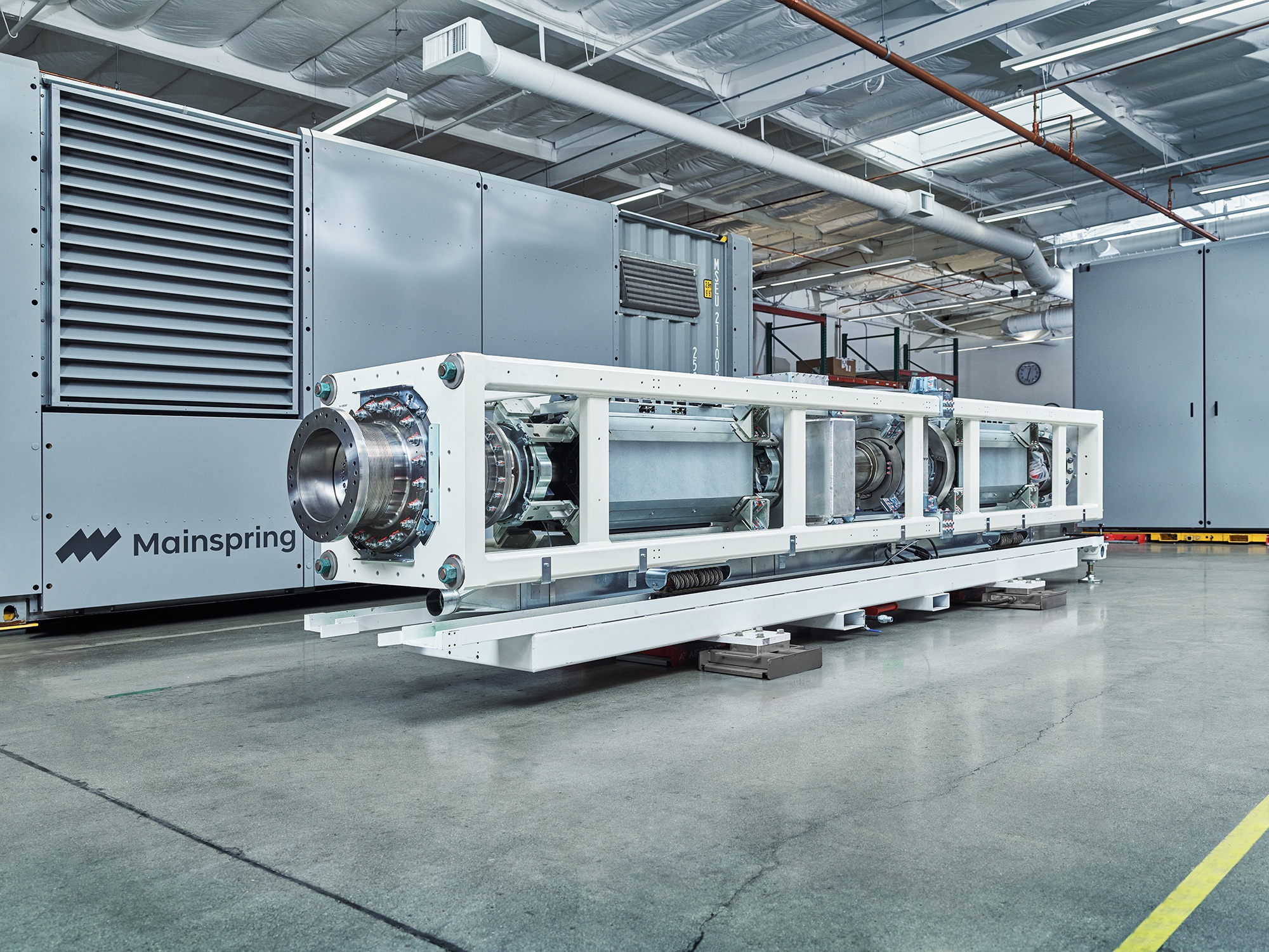 Large industrial linear generator core in a spacious facility with Mainspring equipment in the background.