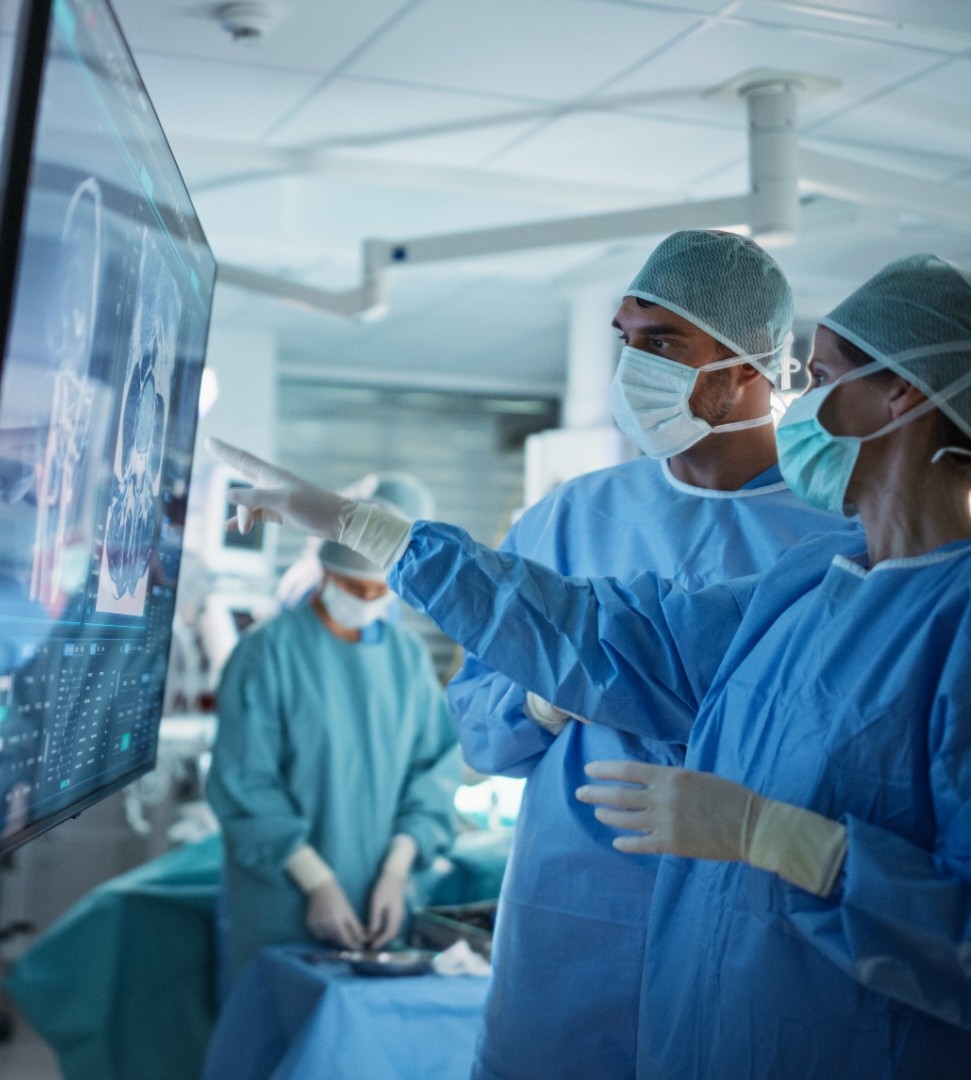 Two surgeons in blue scrubs and masks reviewing brain scans on a large monitor during surgery.