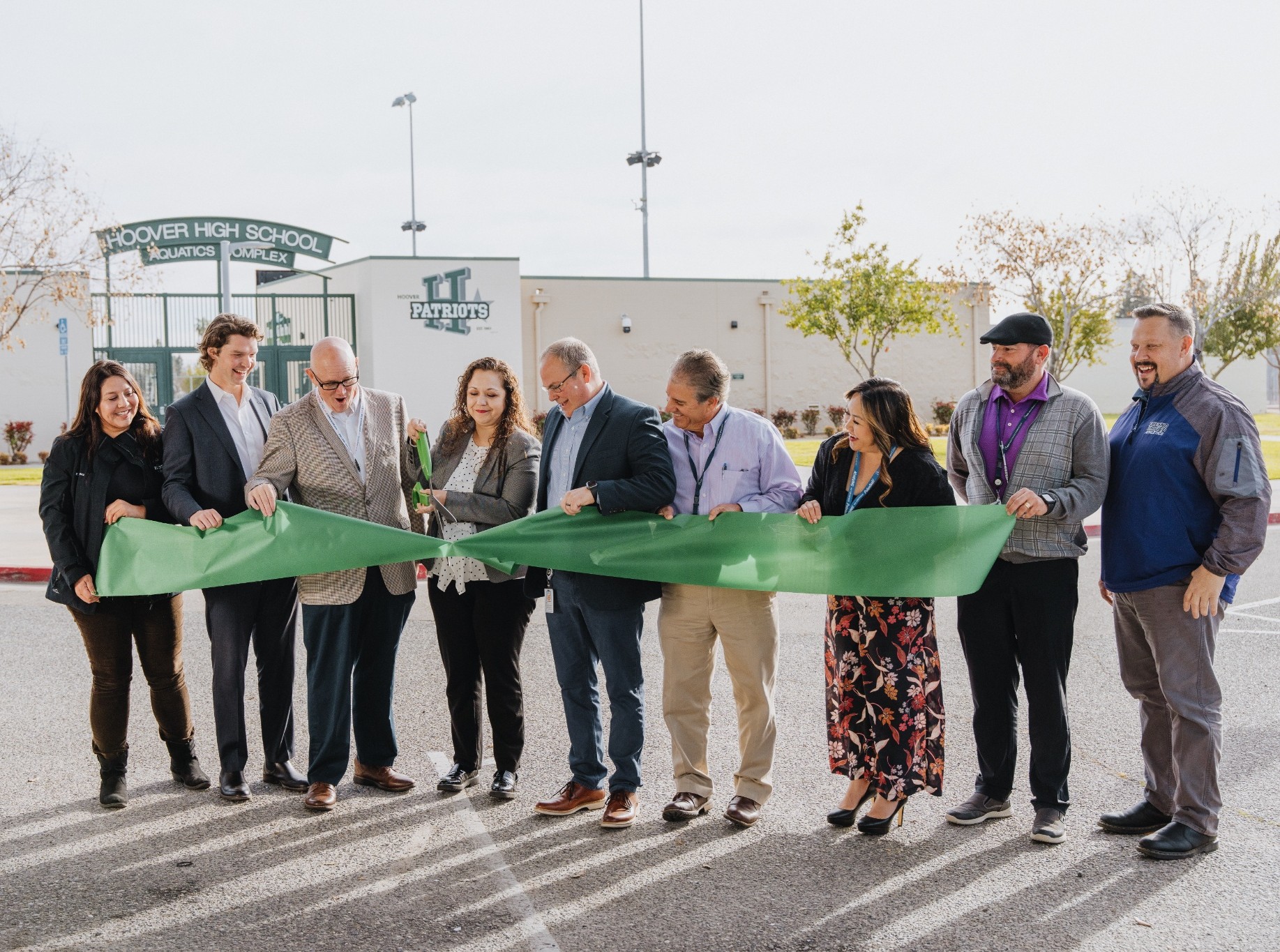 Nine people hold a green ribbon while a man cuts it outside Hoover High School during a ribbon-cutting event.
