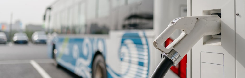 White and blue electric bus charging at a station with a plugged-in charging handle.