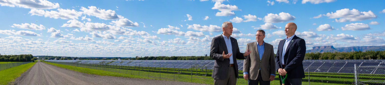 Three men in suits stand near solar panels in a fenced field under a partly cloudy blue sky.