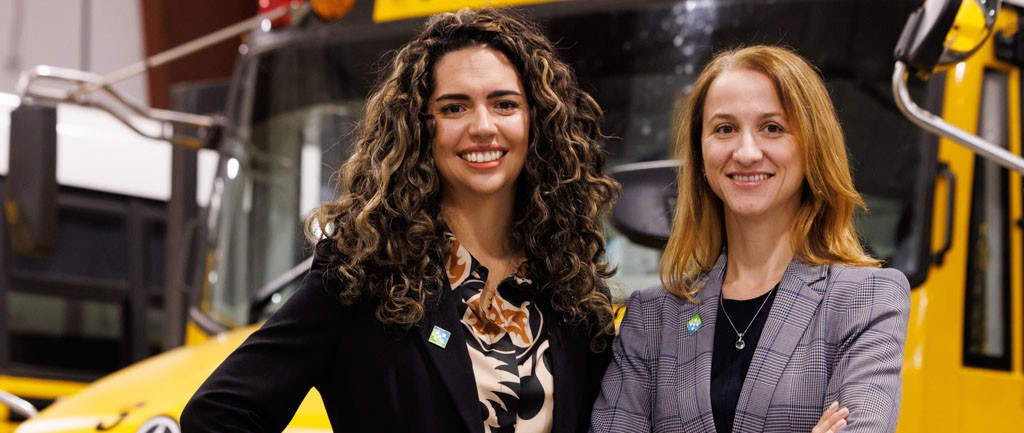 Two women in business attire smiling, standing in front of a yellow bus with arms crossed and relaxed pose.