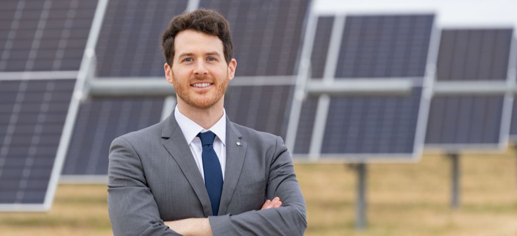 Man in gray suit standing with arms crossed in front of solar panels in an outdoor field.