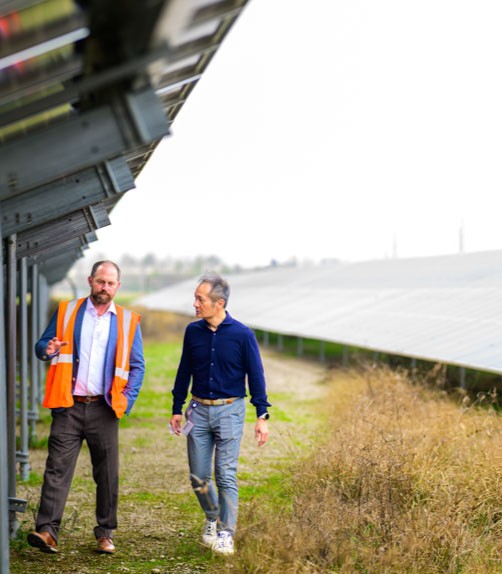 Two men walking and talking between rows of solar panels, one wearing an orange safety vest.