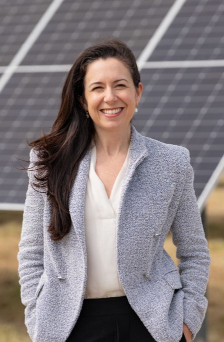 Smiling woman with long hair wearing a gray jacket stands in front of solar panels outdoors.