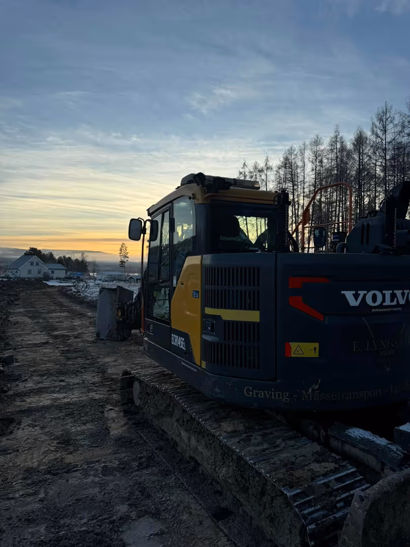 Yellow and black Volvo excavator parked on a dirt path at sunset with bare trees and houses in the background.