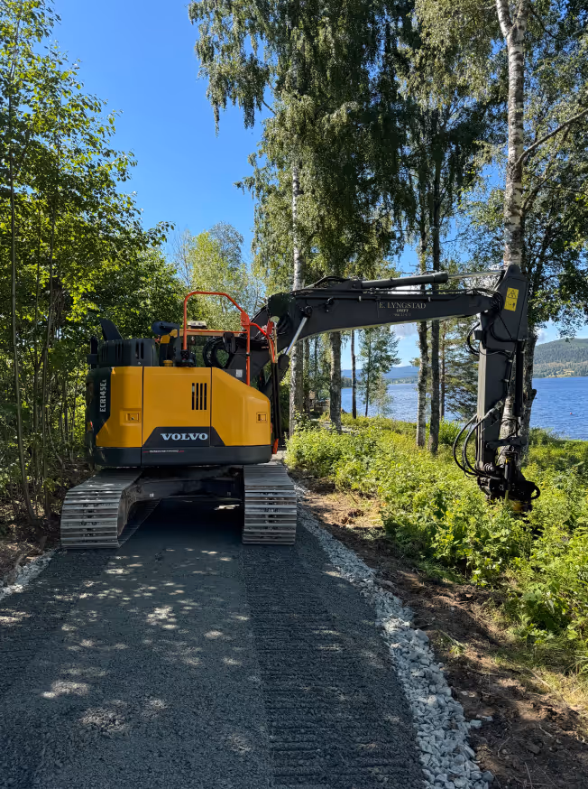 Yellow Volvo excavator with an extended arm clearing bushes on a gravel path near a lake surrounded by tall trees under a clear blue sky.