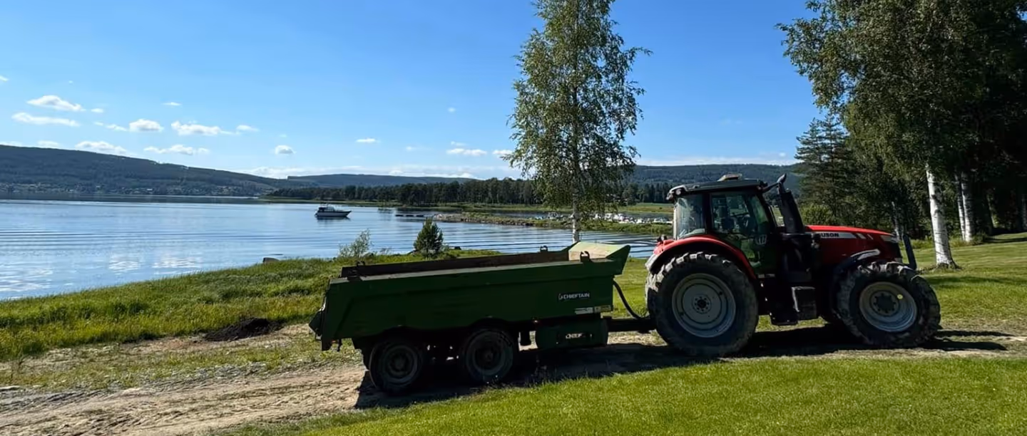 Red tractor attached to a green trailer parked on a grassy area near a calm lake with hills and trees in the background under a blue sky.