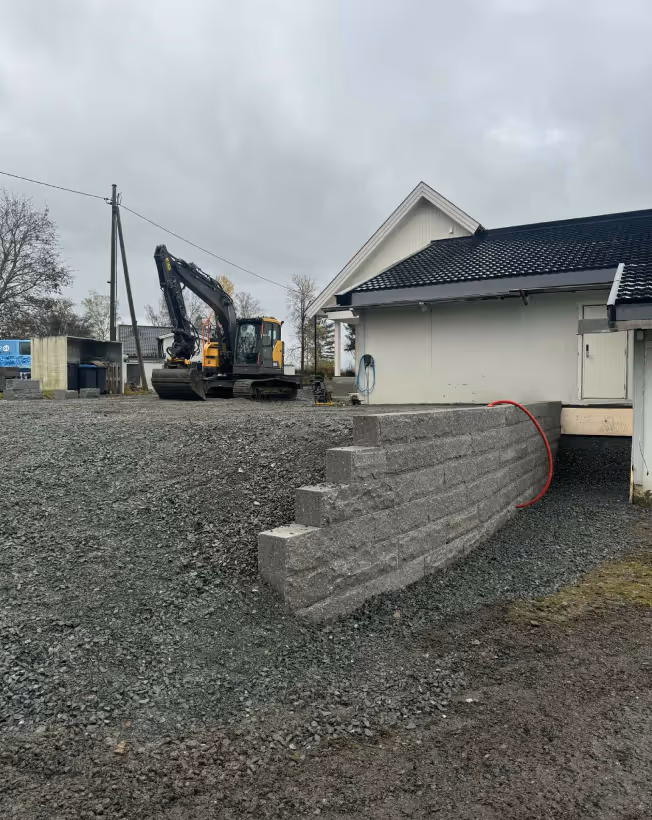 Construction site with a small excavator on gravel beside a white building and a short retaining wall made of concrete blocks.