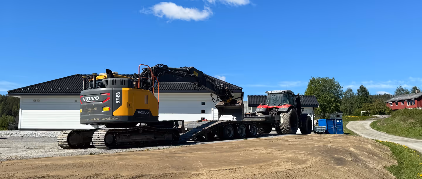 Yellow Volvo excavator parked on a gravel area next to a flatbed trailer attached to a red tractor, with houses and trees under a clear blue sky.