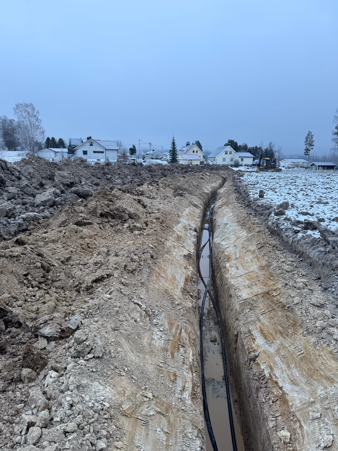 Long trench dug in muddy earth with black cables laid inside, snowy houses and trees in the background under a gray sky.