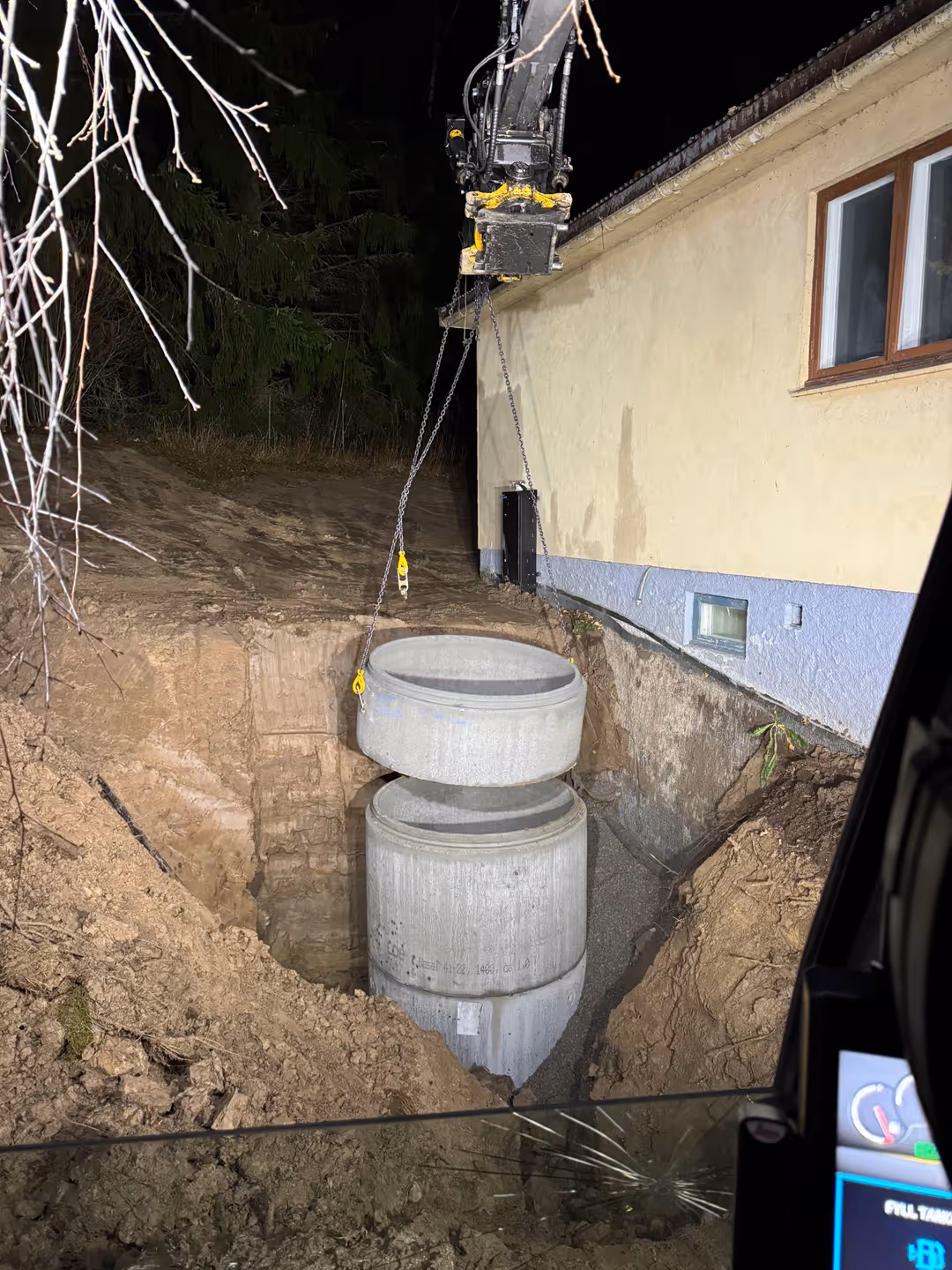 Crane installing large concrete pipes in a deep excavation next to a house at night.