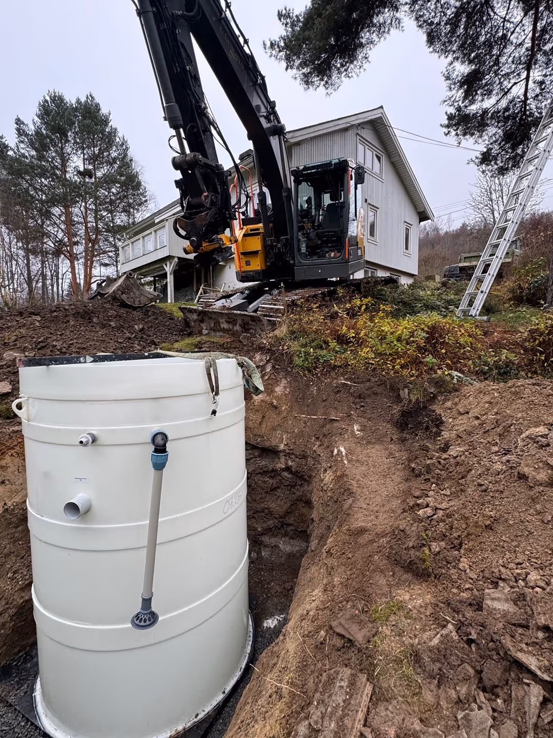 Excavator at a residential construction site with a large cylindrical tank installed in a dug-out hole near a white house.