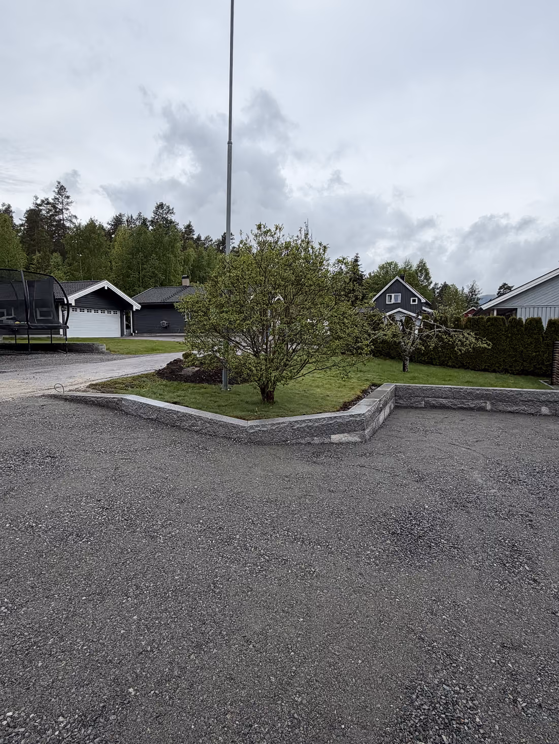 Lav støttemur med busk og gressplener foran boliger under overskyet himmel.