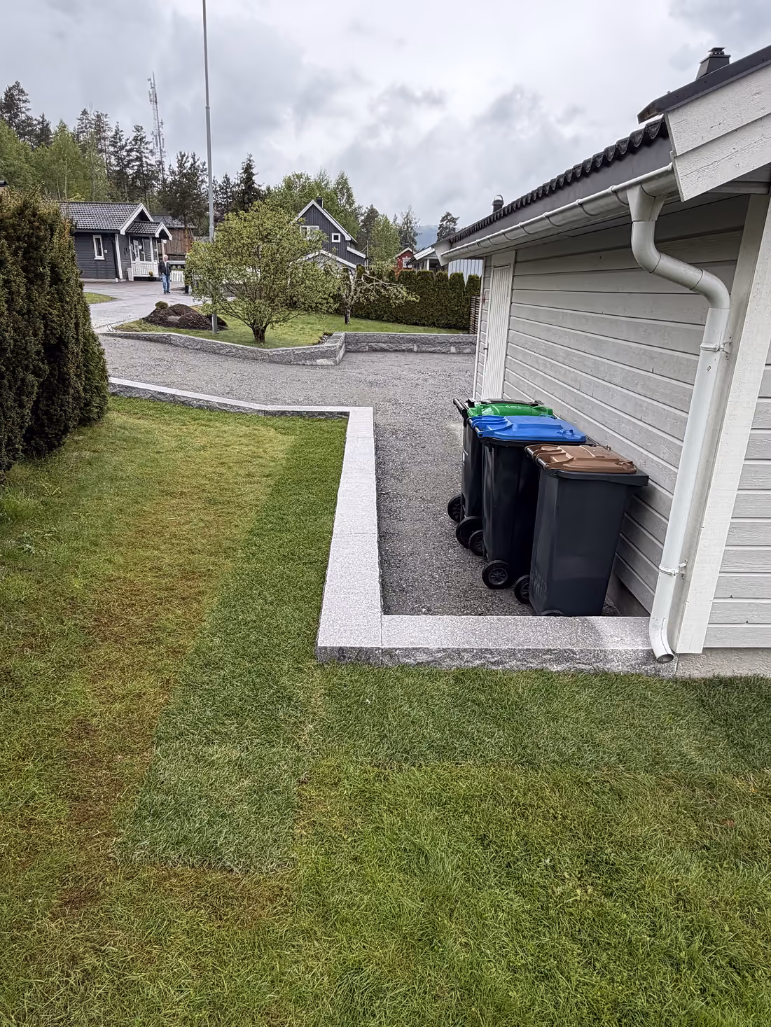 Nytt støttemur i betong ved husvegg med tre søppelkasser og grønt gress rundt.