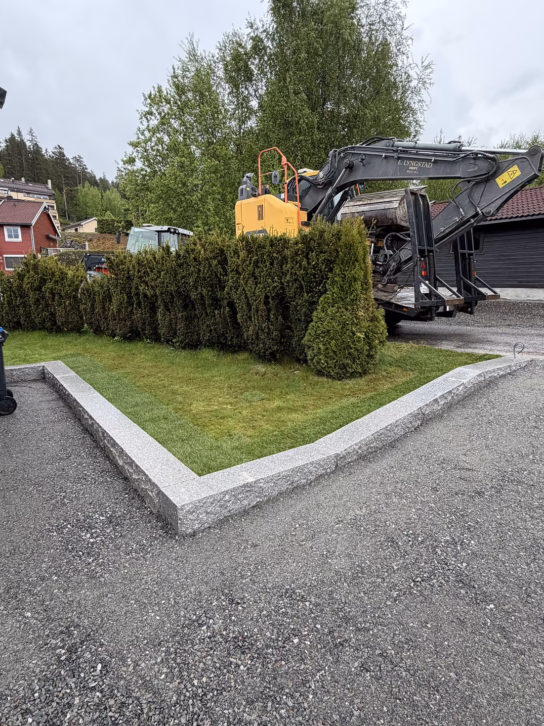 Grå støttemur i granitt avgrenser et hjørne med gress og hekker foran gravemaskin og trær.