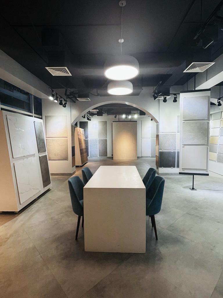 Modern showroom with a white table and four blue chairs in the center, surrounded by large tile samples displayed on walls and stands.