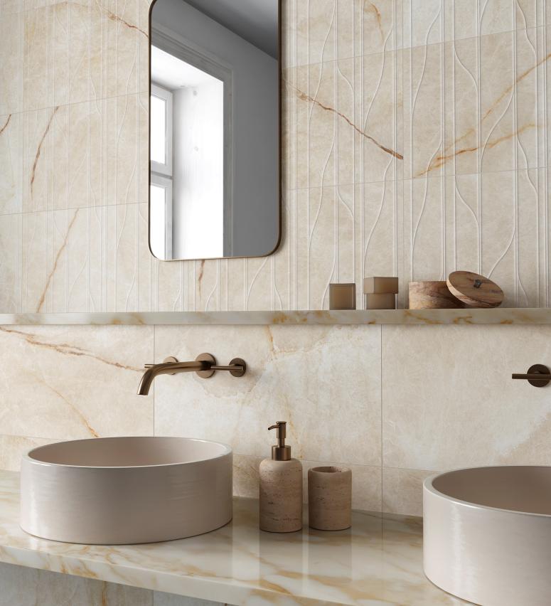 Modern bathroom countertop with two round beige sinks, wall-mounted bronze faucets, and a vertical rectangular mirror reflecting a window.
