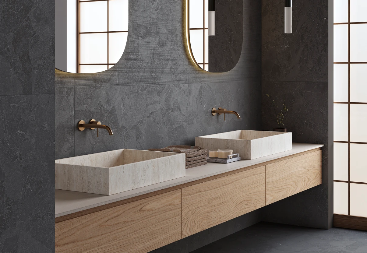 Modern bathroom with dual rectangular stone sinks, gold wall-mounted faucets, dark stone walls, wooden vanity, and backlit mirrors.