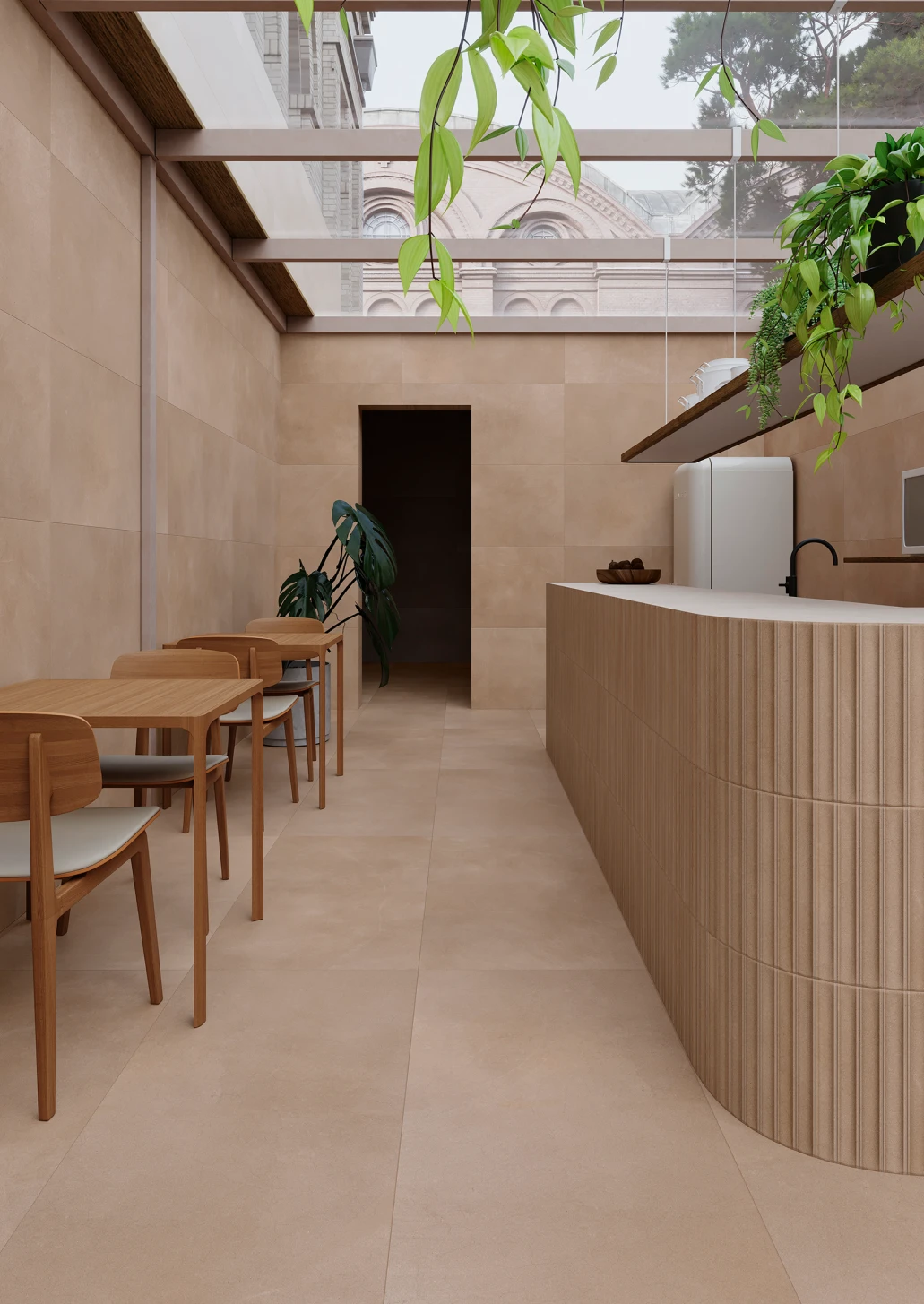 Minimalist cafe interior with beige porcelain tile walls and floors, wooden tables and chairs, a rounded wooden counter, and hanging plants with a glass ceiling showing a building outside.