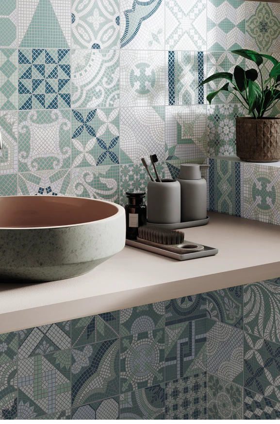 Bathroom countertop with a round vessel sink, gray soap dispenser and toothbrush holder, a small bottle, brush, and a potted plant against a multi-patterned blue and green tiled wall.