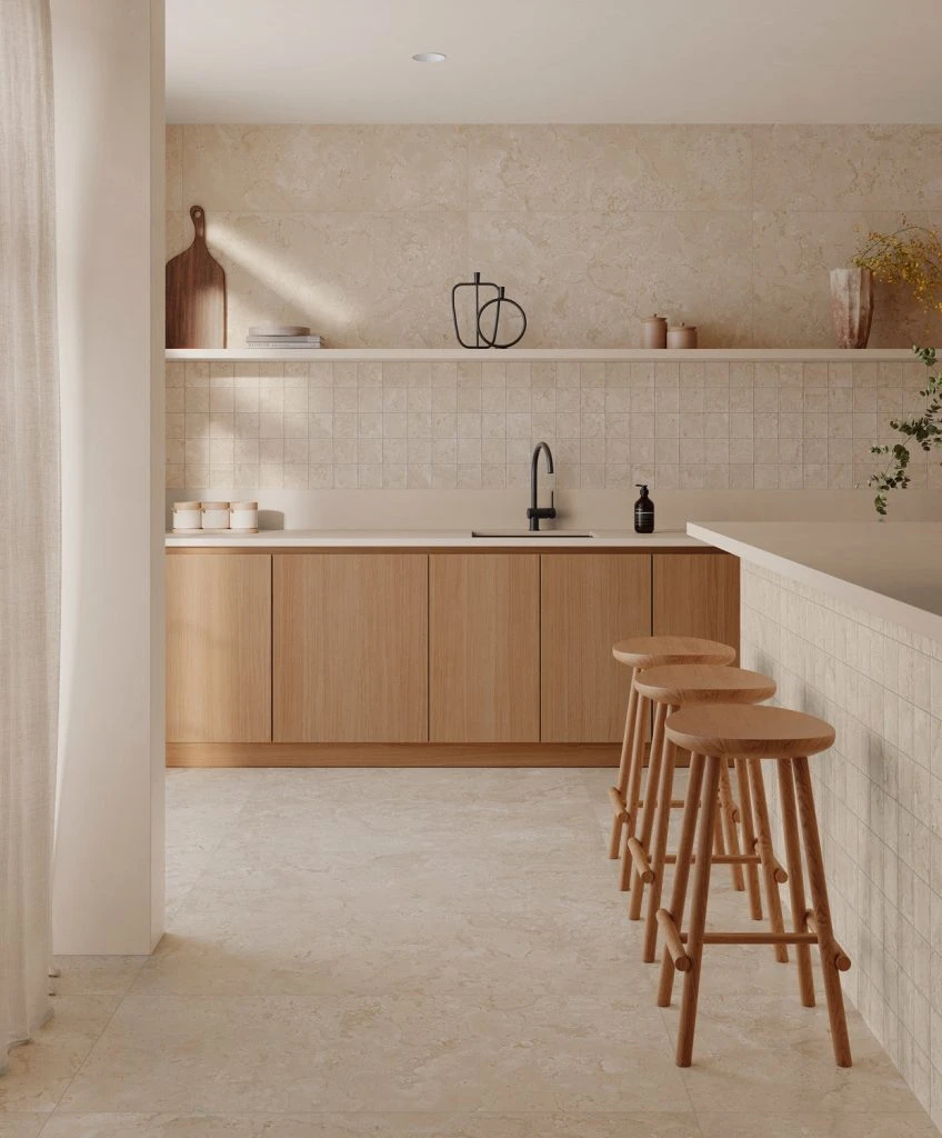 Minimalist kitchen with light beige stone tiles, wooden cabinets, three wooden stools, and a black faucet.