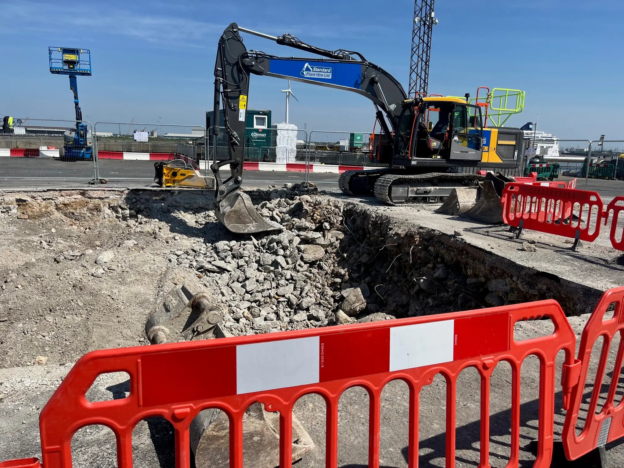 Excavator digging a construction site surrounded by red and white safety barriers under a clear blue sky.