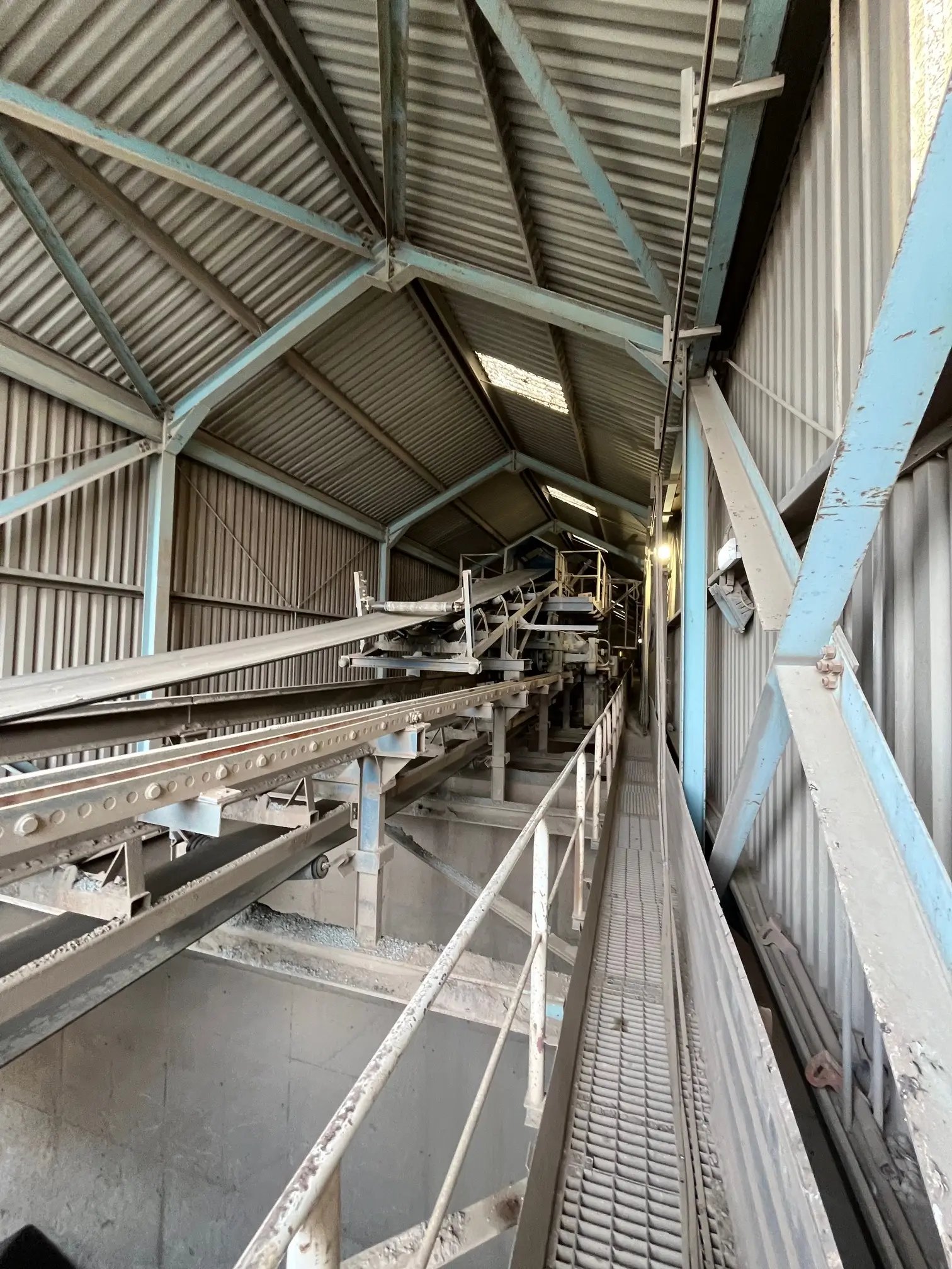 Interior of an industrial facility with metal conveyor belts and a grated walkway under a corrugated metal roof.
