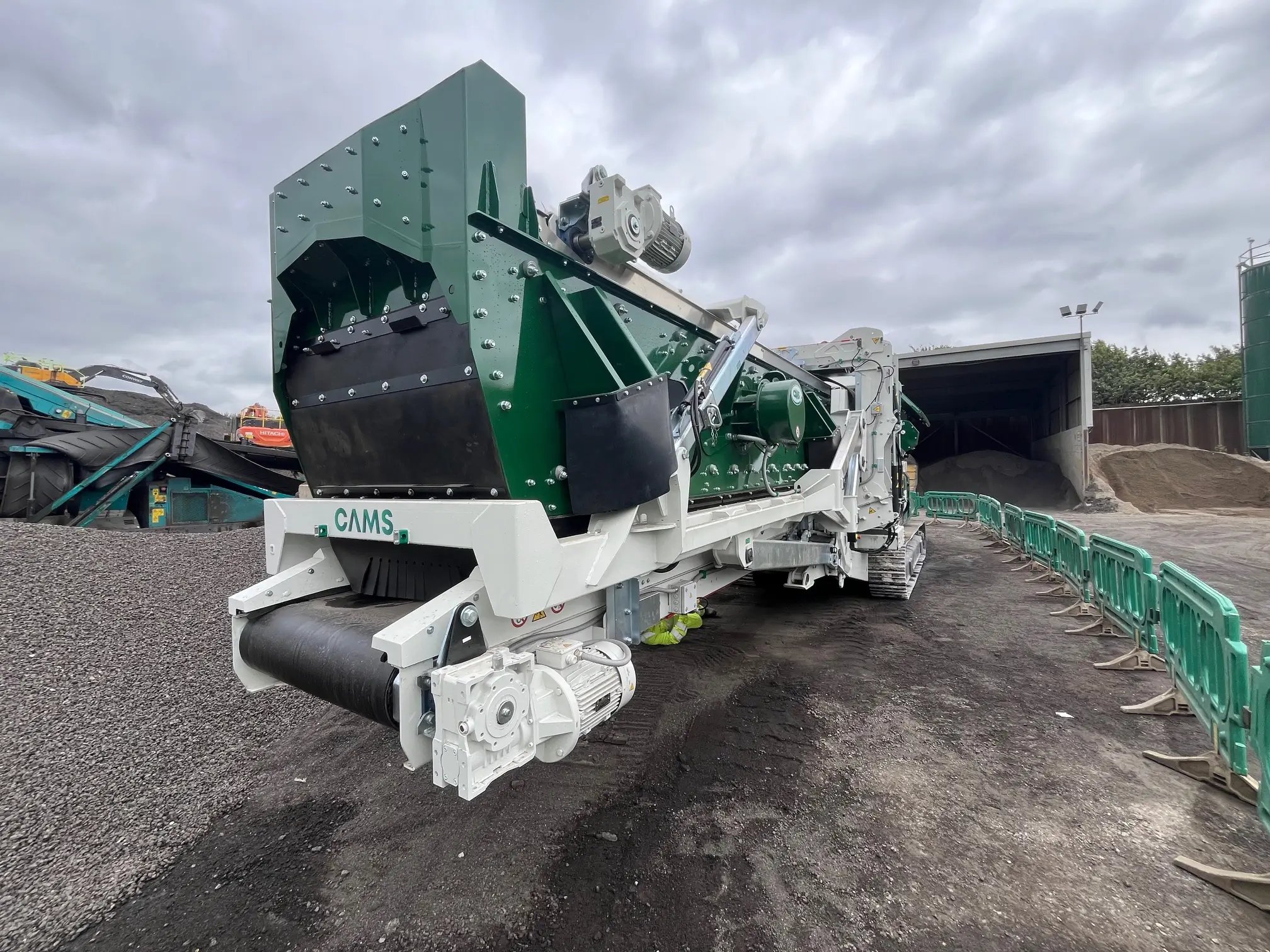 Large industrial green and white CAMS machine with conveyor belt on a gravel surface under a cloudy sky.