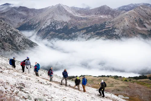 Group of hikers walking on a snowy mountain slope above a cloud-filled valley with rugged peaks in the background.