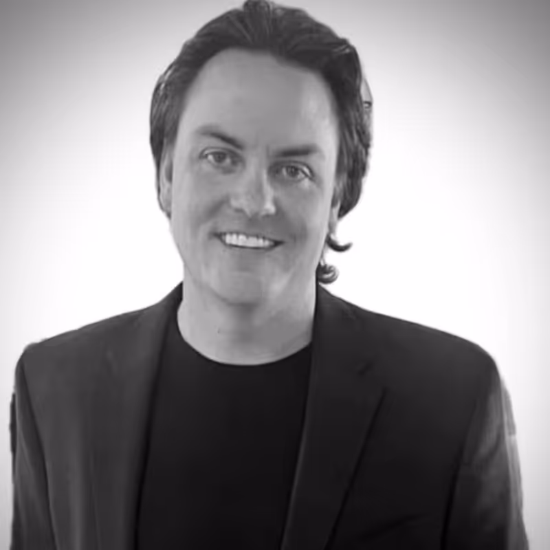 Portrait of a man with dark hair smiling, wearing a black jacket over a black shirt, on a plain light background.