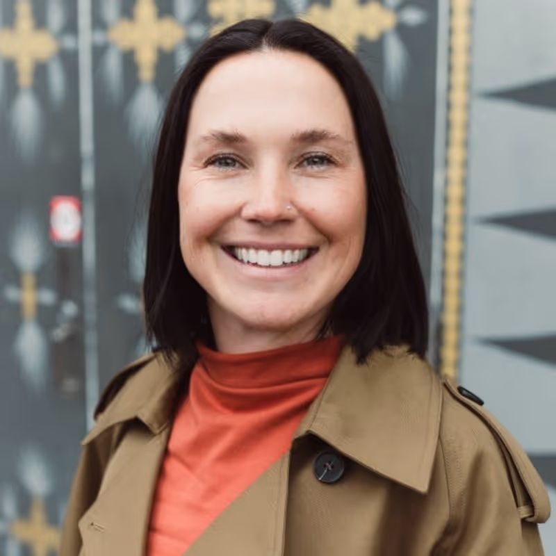 Smiling woman with shoulder-length dark hair wearing a tan coat and rust-colored top standing outdoors.