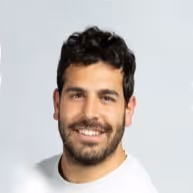 Smiling man with dark curly hair, beard, and mustache wearing a white shirt against a light gray background.