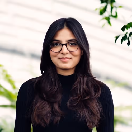 Portrait of a woman with long dark hair and black glasses wearing a black top, with blurred greenery and white background.