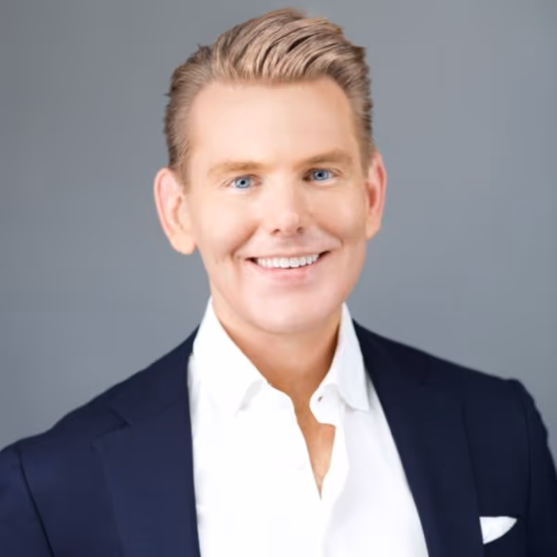 Smiling man with light hair and blue eyes wearing a white shirt and dark blazer against a gray background.
