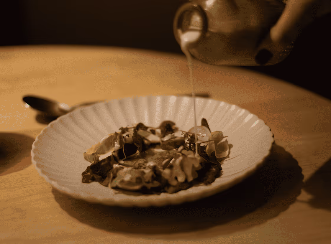 Mano vertiendo una salsa cremosa sobre un plato blanco con setas y hierbas frescas.