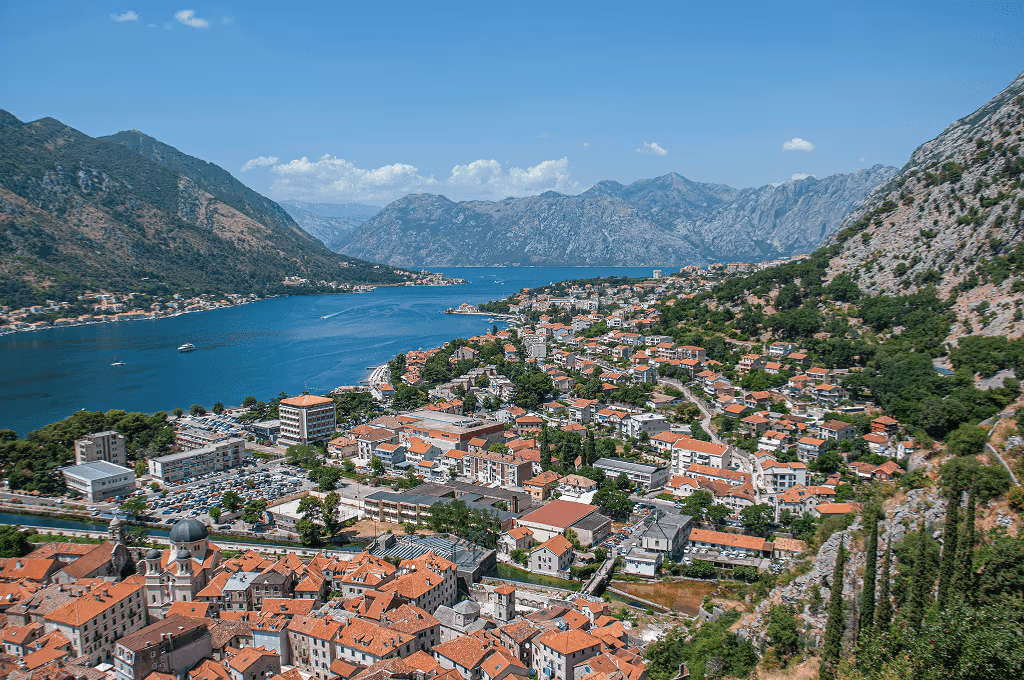 Aerial view of a coastal town with red-roofed buildings, surrounded by mountains and a deep blue bay under a clear sky.