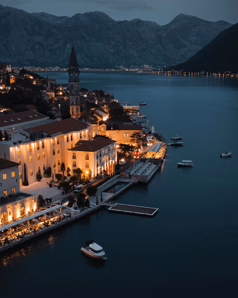 Evening view of coastal town with illuminated buildings, boats on calm water, and mountains in the background.