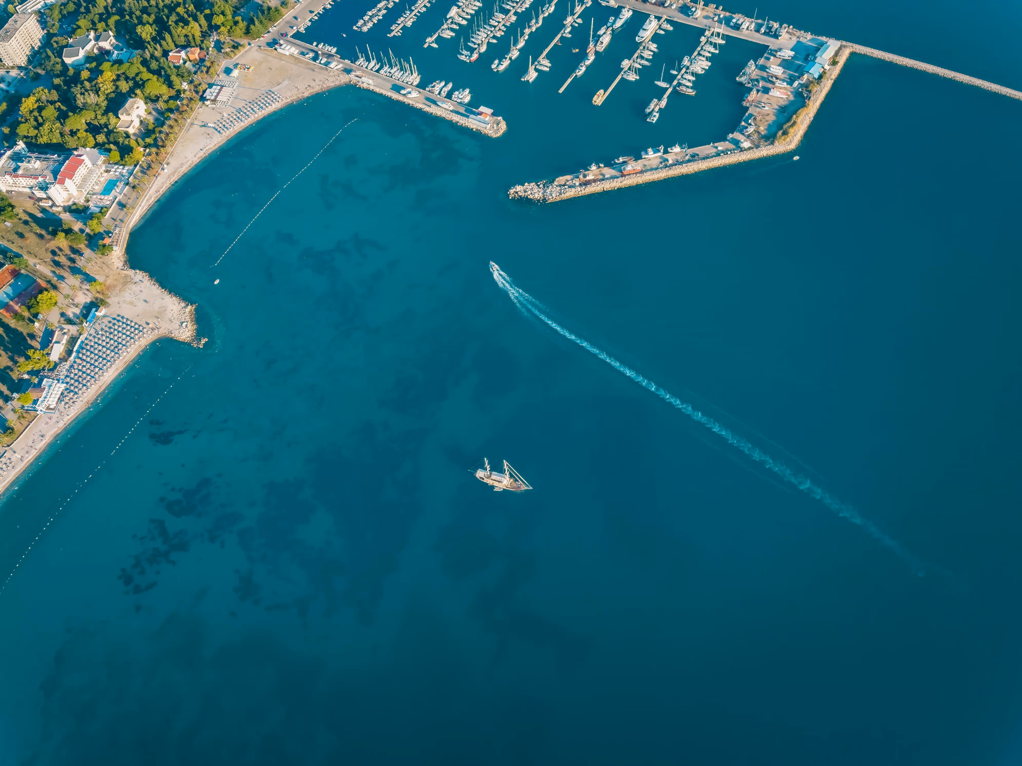 Aerial view of a marina with moored boats, a sandy beach lined with umbrellas, a green park, and a boat leaving a white wake in blue water.
