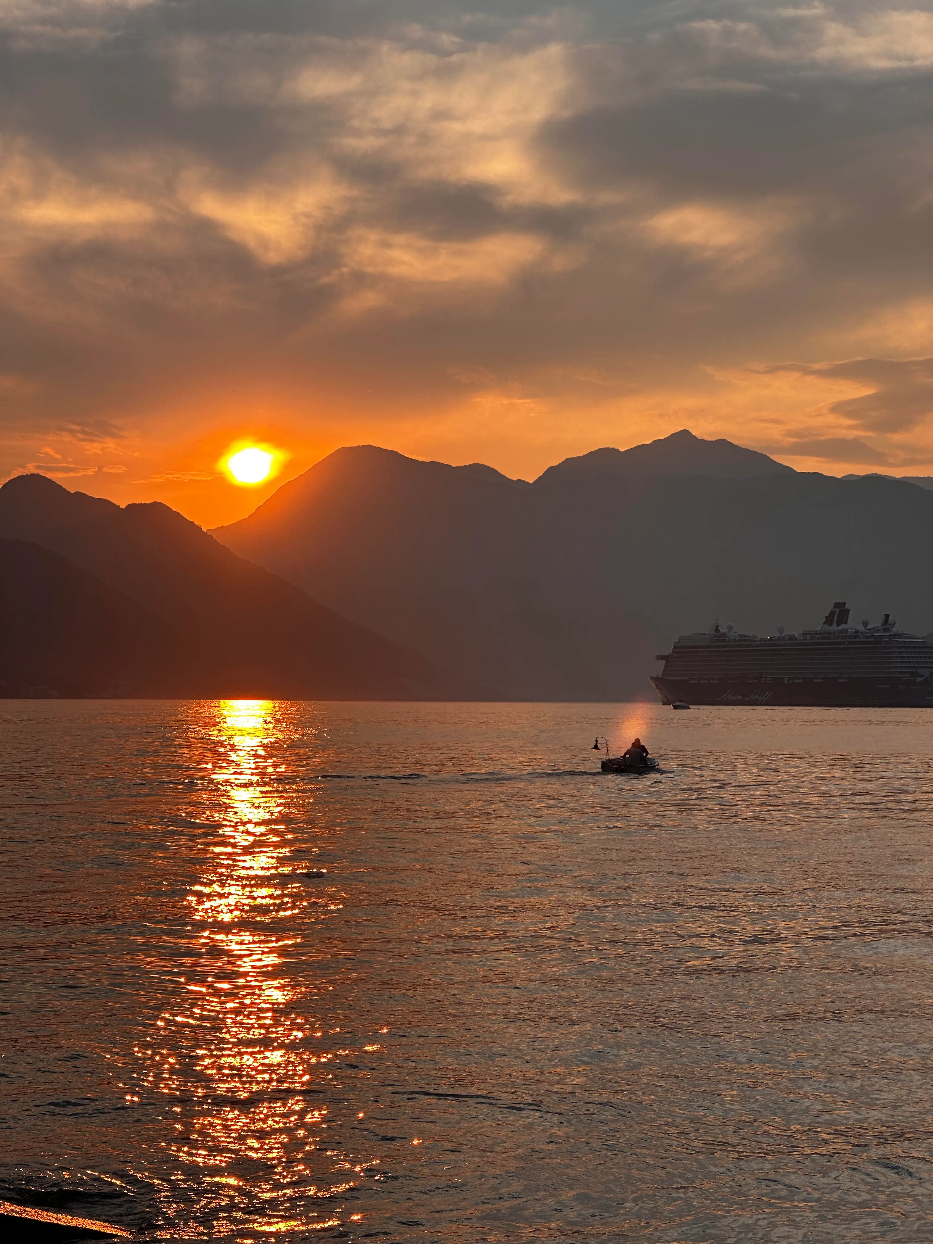 A small fishing boat alongside a cruise liner at sunset on Kotor Bay, mountains silhouetted behind, warm evening light