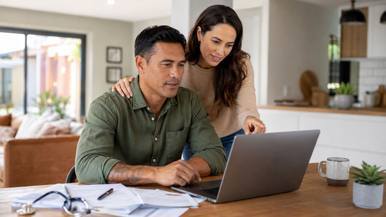 Alt text: Veteran seated at a laptop with a supportive partner beside him, reviewing DVA cancer treatment paperwork in a bright home setting.