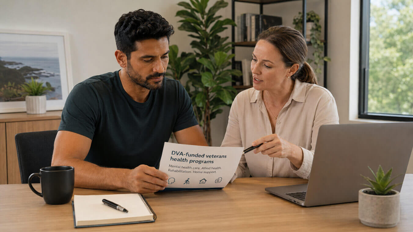 Australian veteran reviewing DVA health program options with a female consultant in a modern office, with paperwork, a laptop and care resources on the desk.