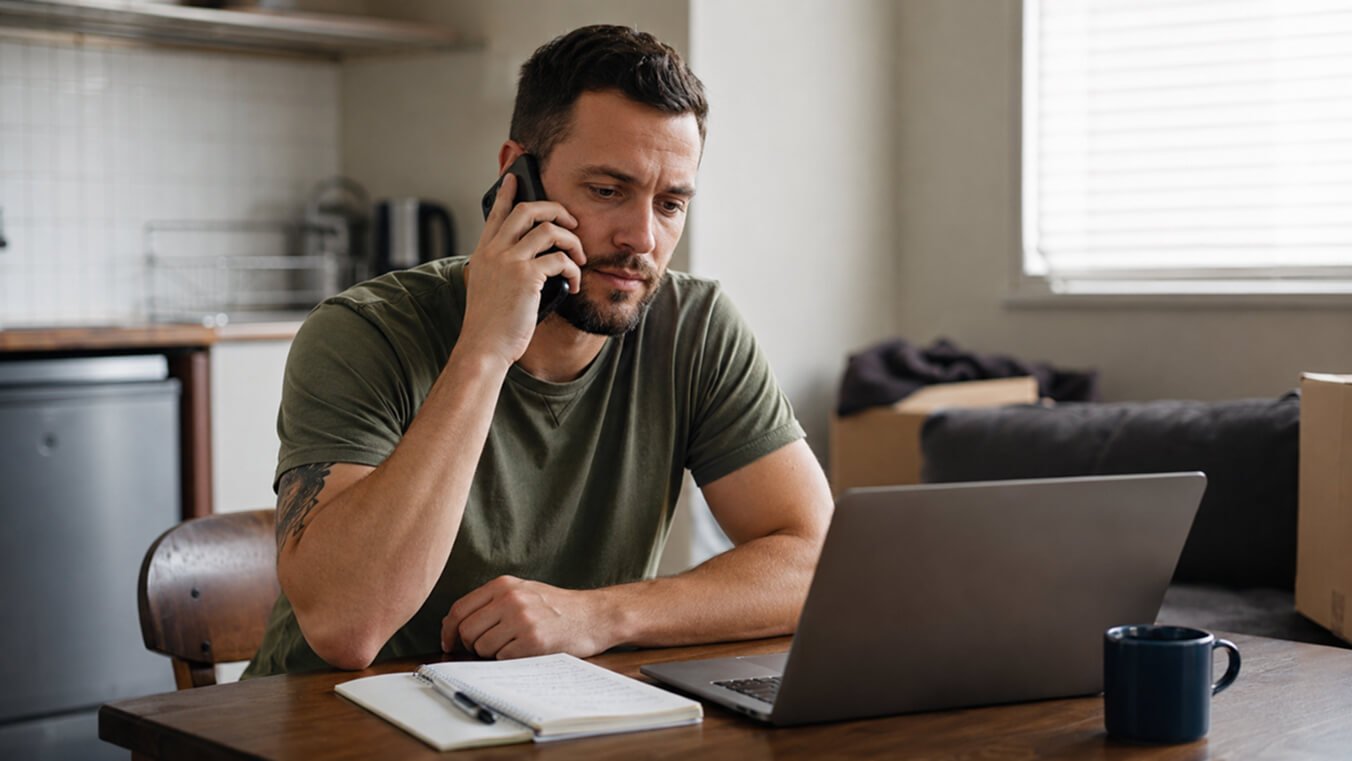 Young veteran sits at a table in a modest home, speaking on the phone while looking at a laptop, with a notebook and coffee mug nearby as he seeks emergency housing support.