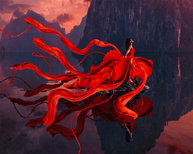 Woman on a reflective mountainous background wearing a red gown with a flowing multi-layered train in the wind taken by Kristina Makeeva