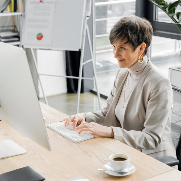 Middle-aged woman with short hair and hoop earrings typing on a keyboard at a desk with a cup of coffee.