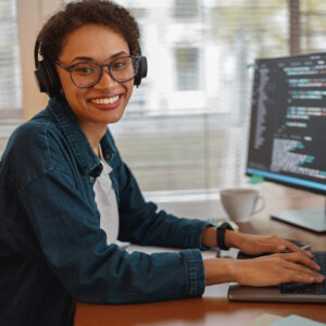 Smiling woman wearing glasses and headphones typing on a keyboard in front of a computer screen displaying code.