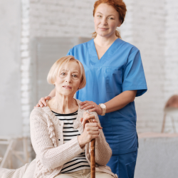 Smiling nurse in blue scrubs standing behind elderly woman holding a cane, offering support.