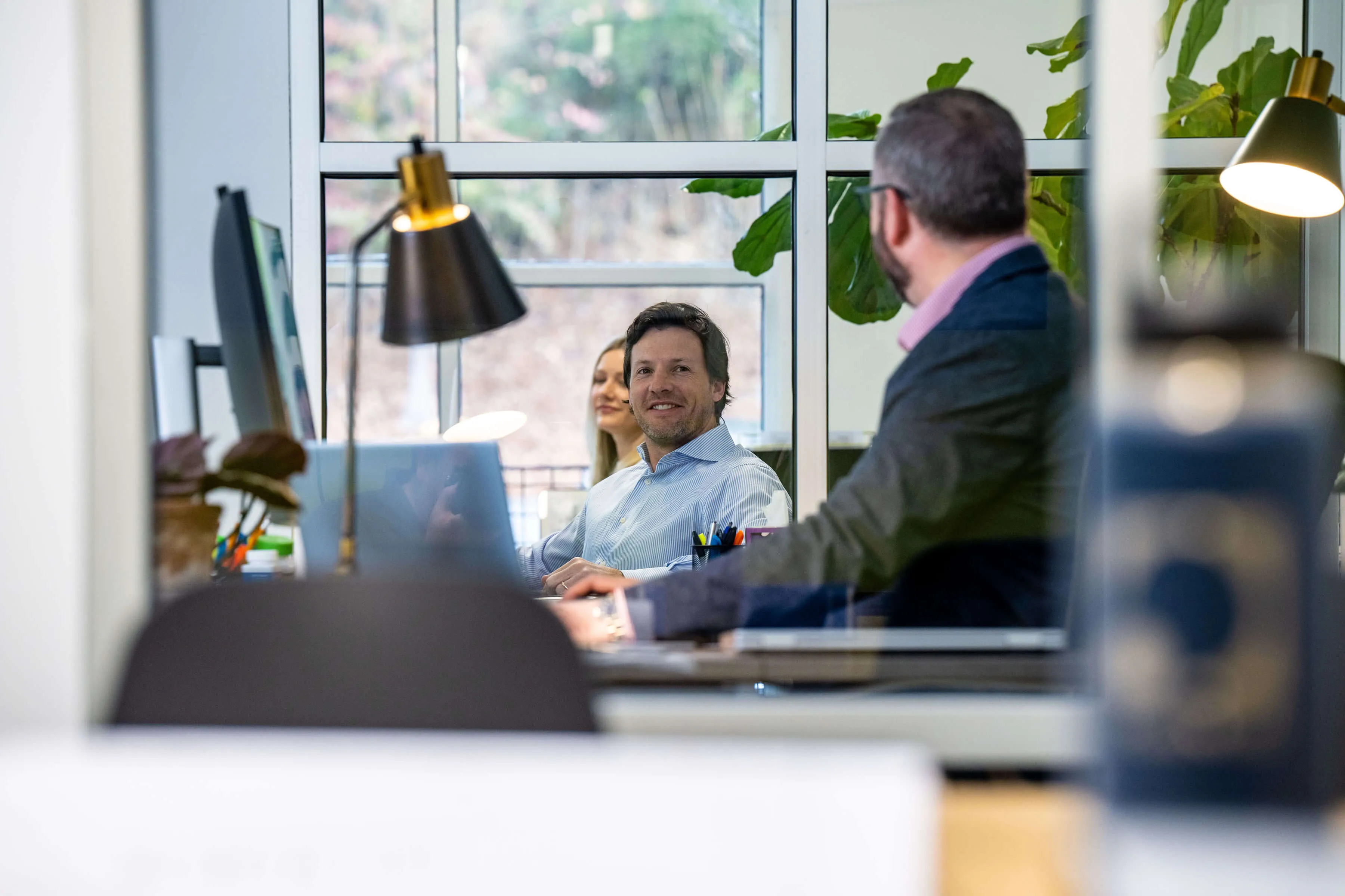 Two men and woman working and talking in an office with large windows and desk lamps.