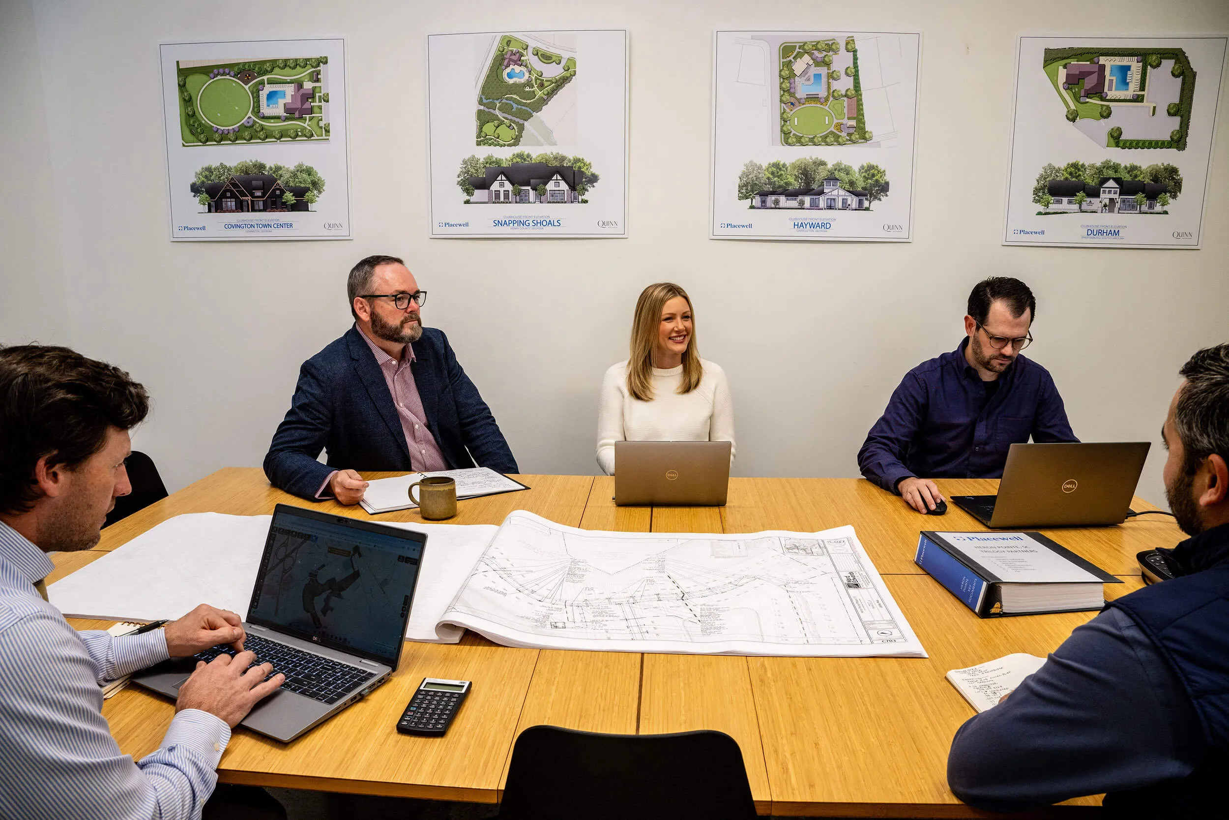 Five people around a table with laptops, papers, and architectural plans in a meeting room.