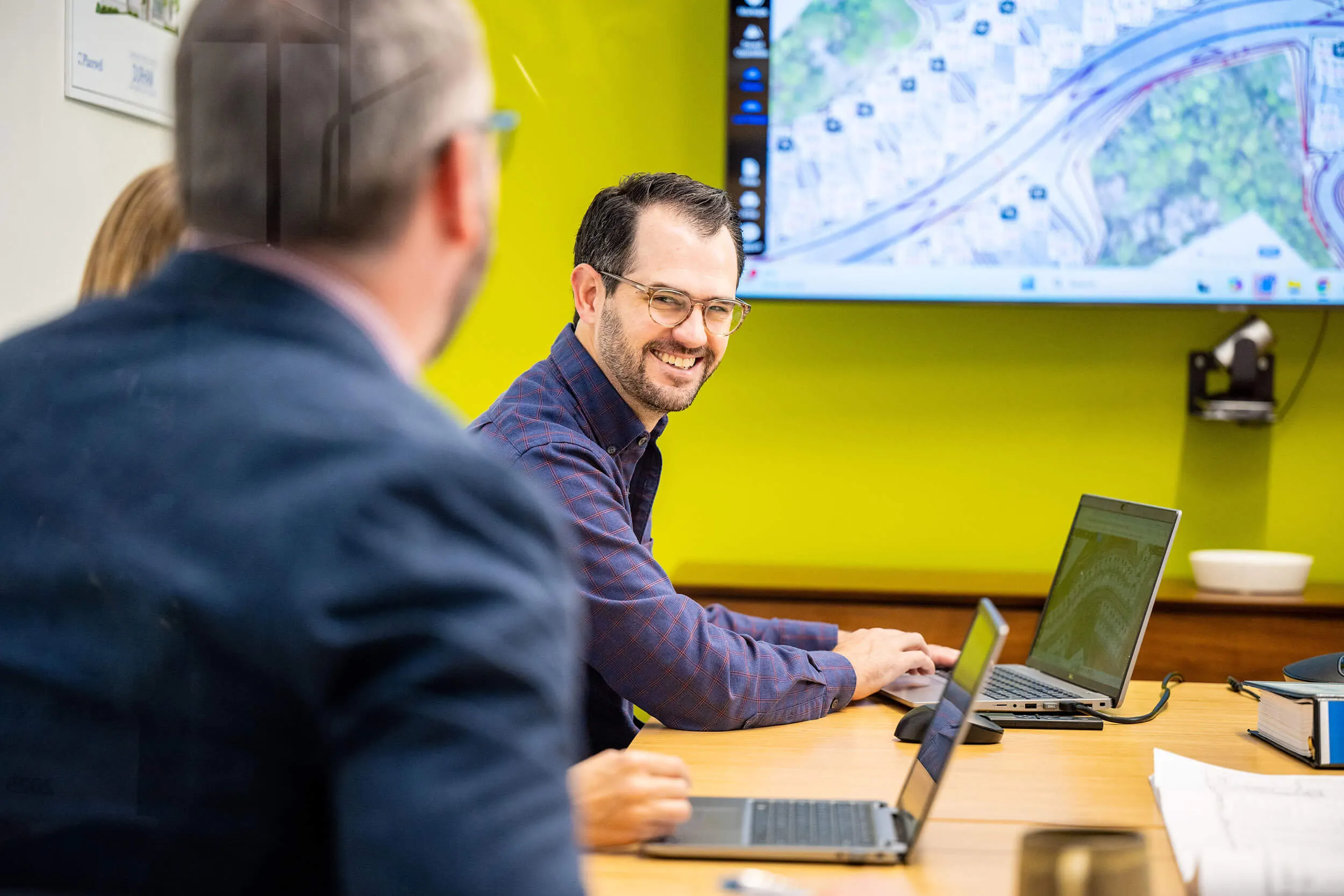 Man smiling at a meeting table with laptops and map displayed on screen in a bright office.